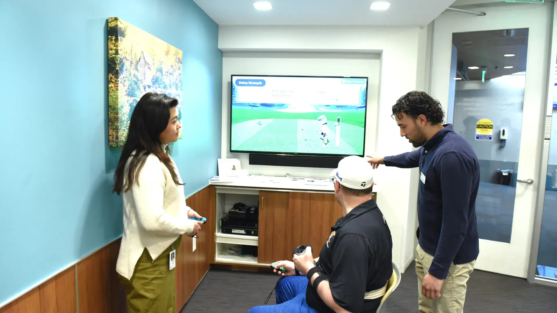 A man sits in a chair with a gaming controller in his hand looking at a television screen with a golf video game while another man points at the screen and a woman looks on