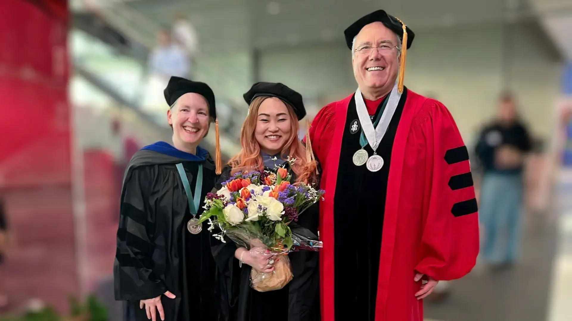 A woman holding flowers wearing a graduation robe stands between a man to her left with a red graduation robe and two medals and a woman in a black graduation robe with one medal
