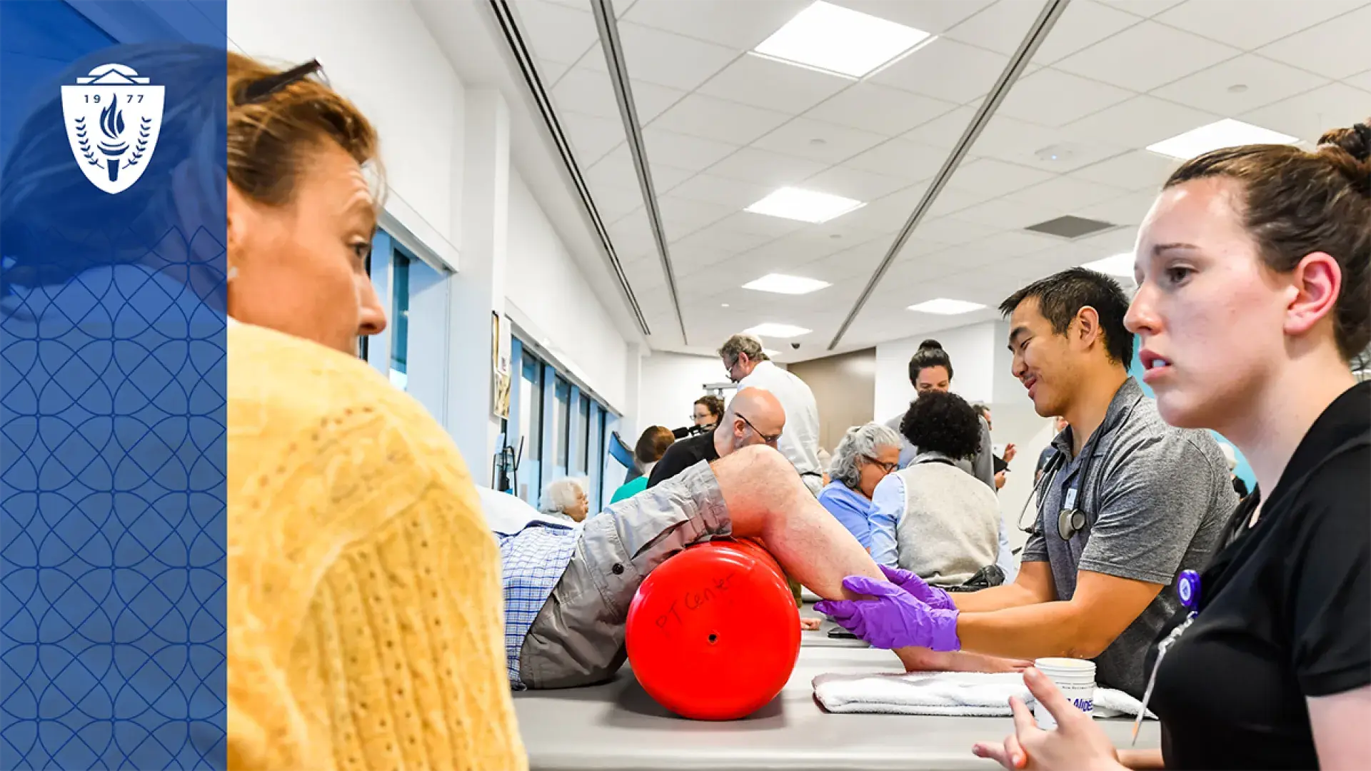 a man helps an older man roll his legs on a red foam tube on a treatment table while two women talk in the foreground