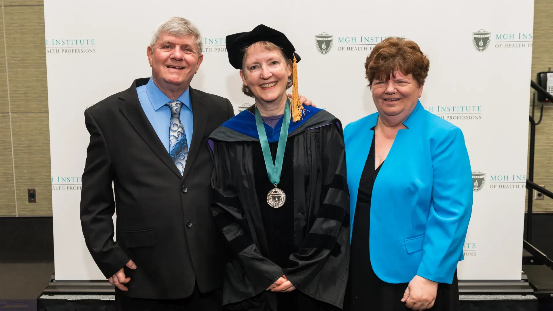 A woman wearing a black robe and a medal around her next poses with a man to her right and a woman to her left