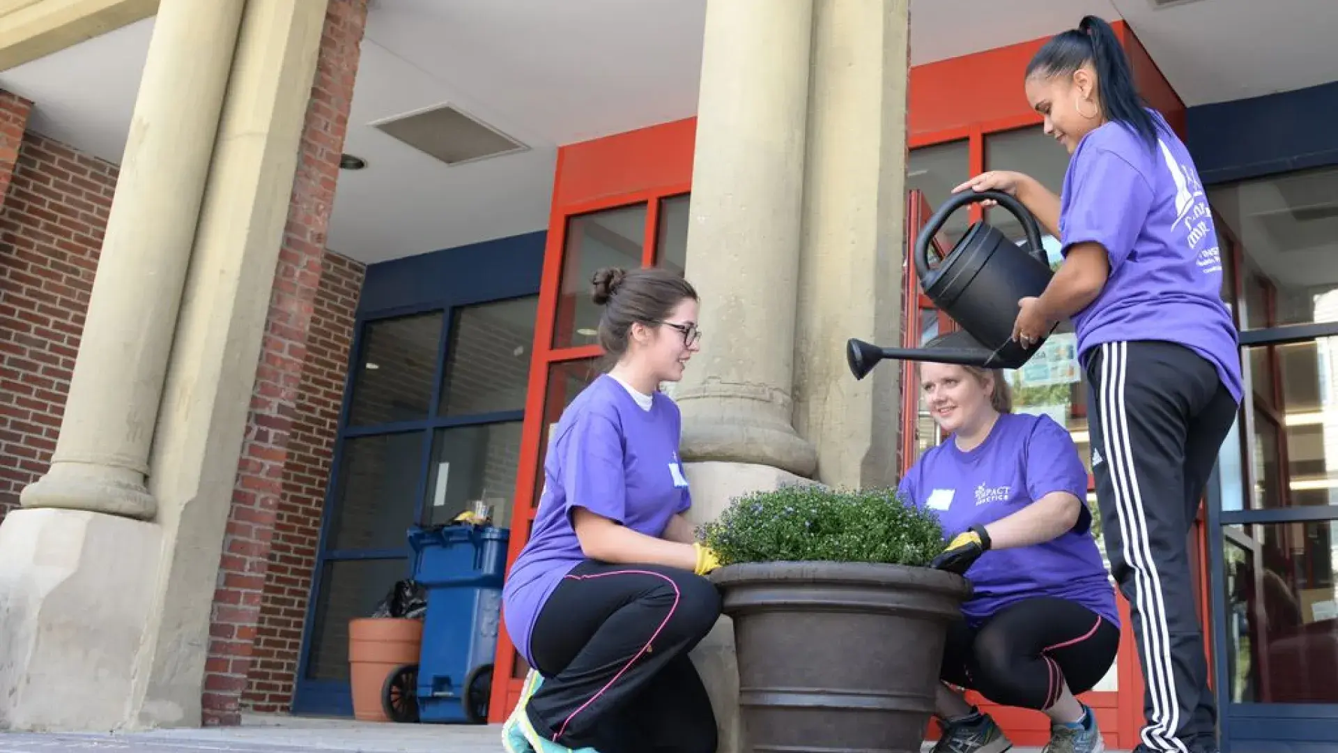 Two women kneel near a flowerpot while a third woman waters the plant
