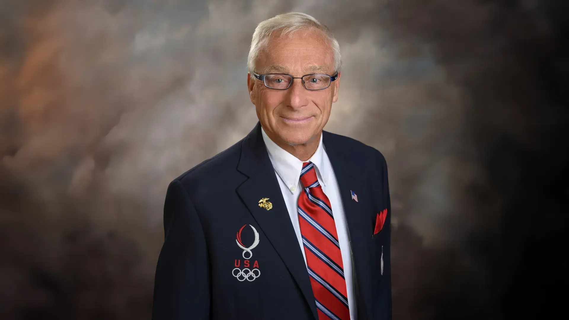 george wears a navy blazer with white Olympic rings on it and a red white and blue striped tie