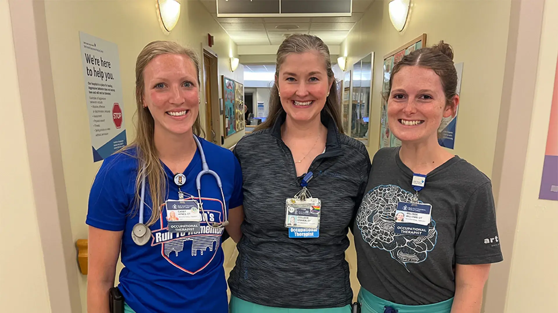 three women in matching scrub bottoms stand in a clinical hallway arm and arm 