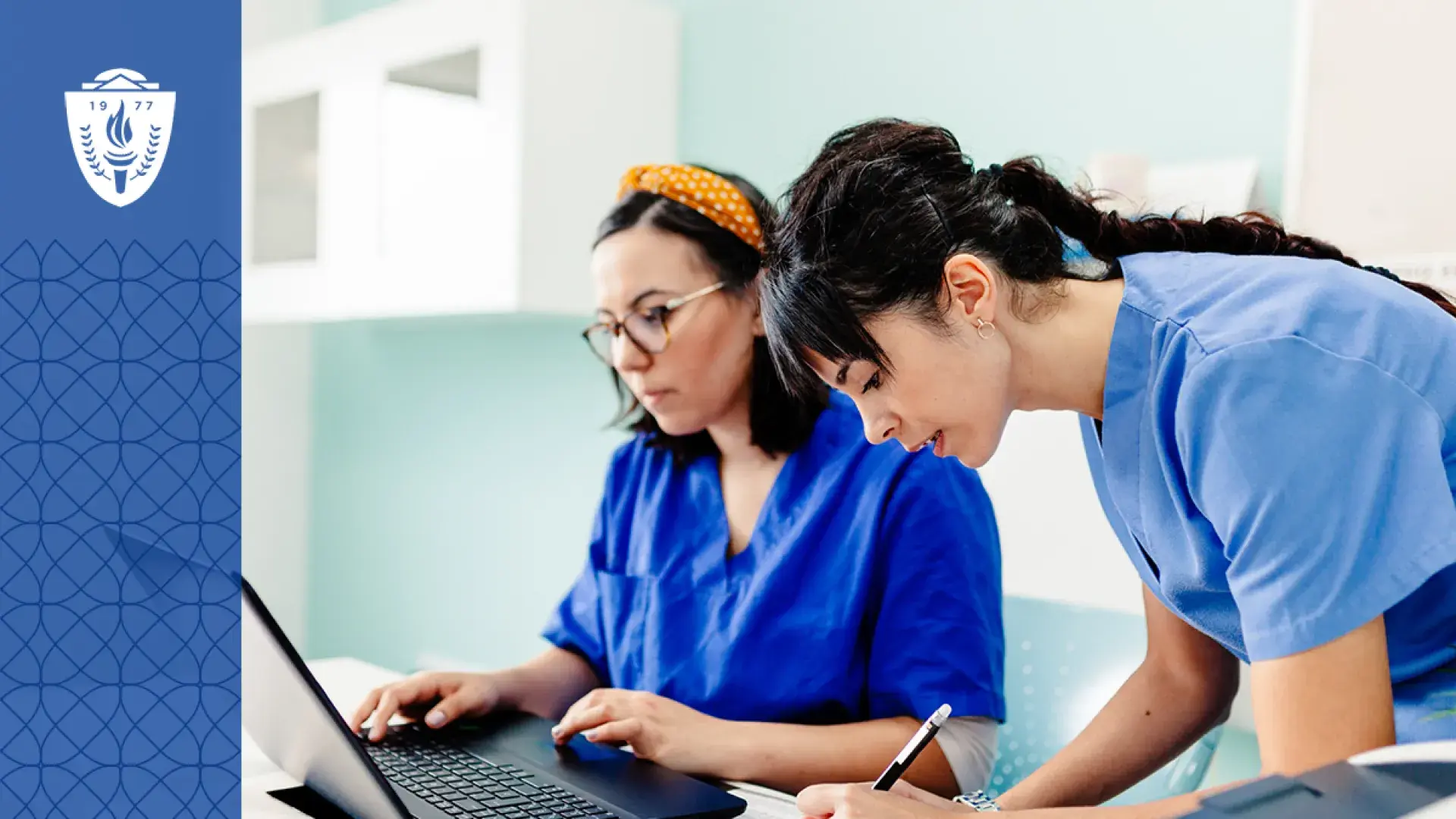 two women in scrubs work at a table with a laptop and pencil