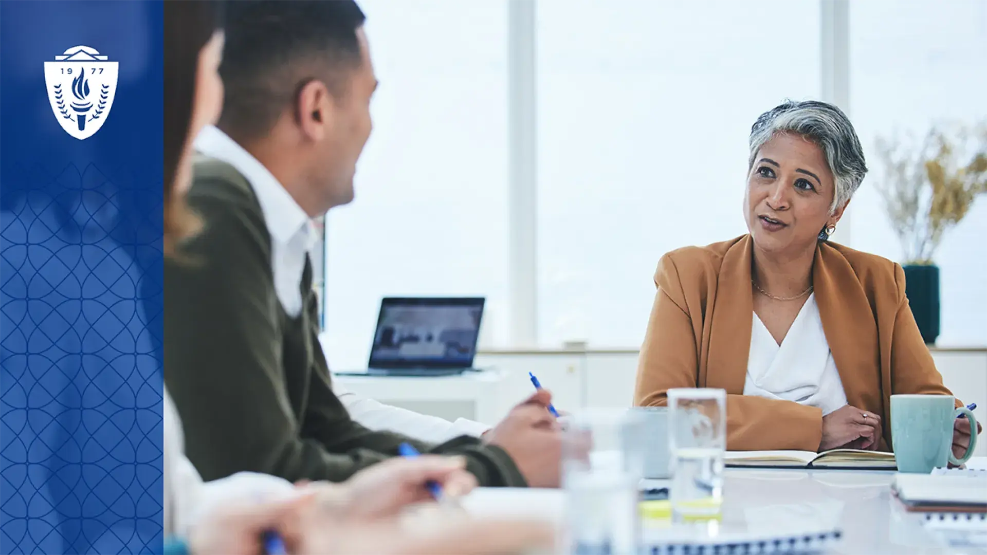 a woman with short grey hair sits at a conference table and talks to people in collared shirts