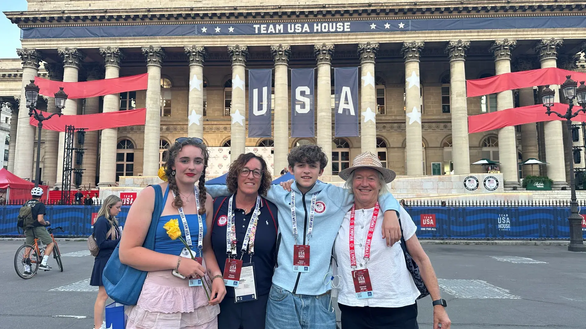 Four people stand in front of a large building with columns that has a USA banner and says Team USA House across the top of the building