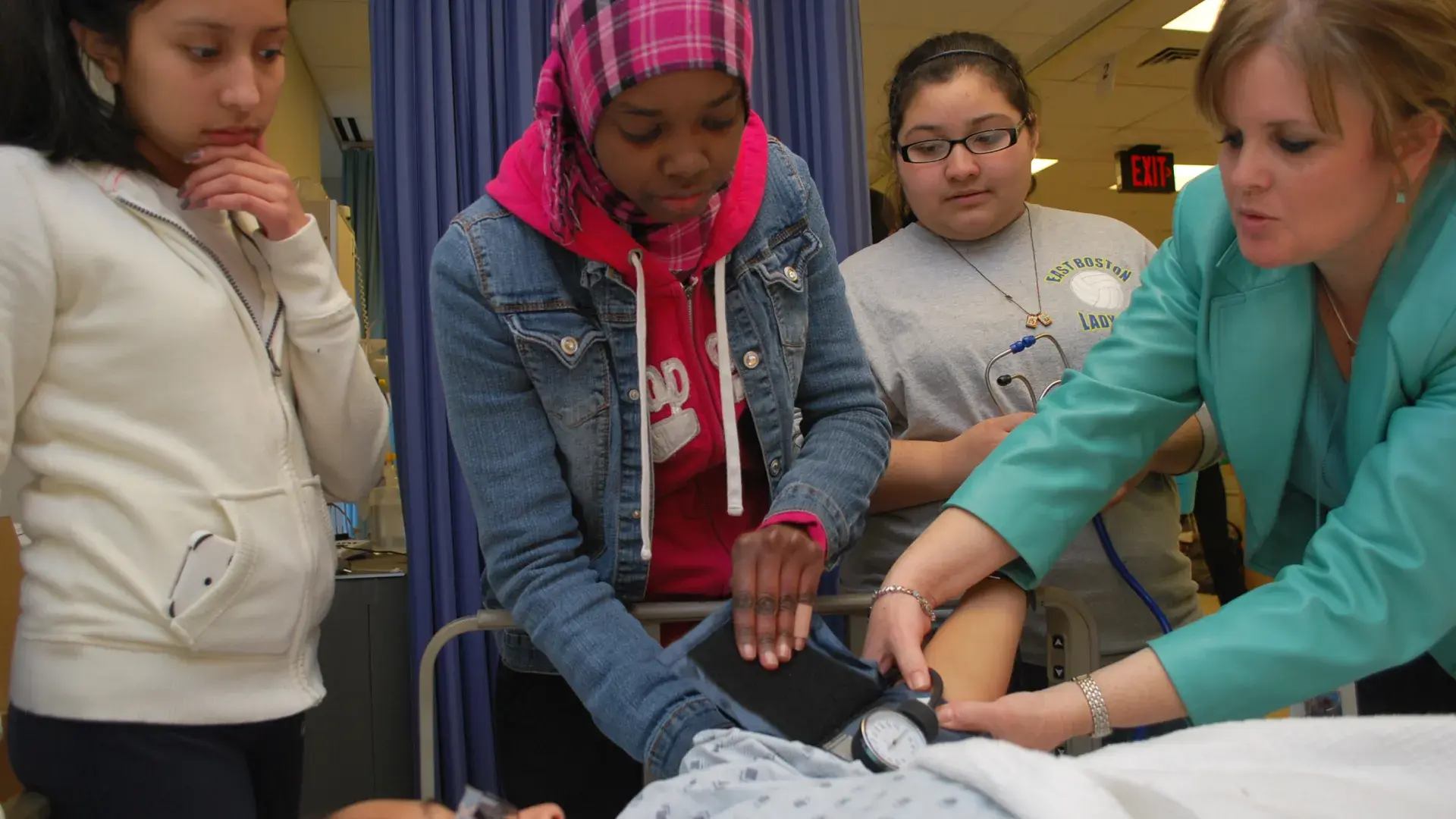 A woman in a hospital setting demonstrates taking a patient's blood pressure to three girls