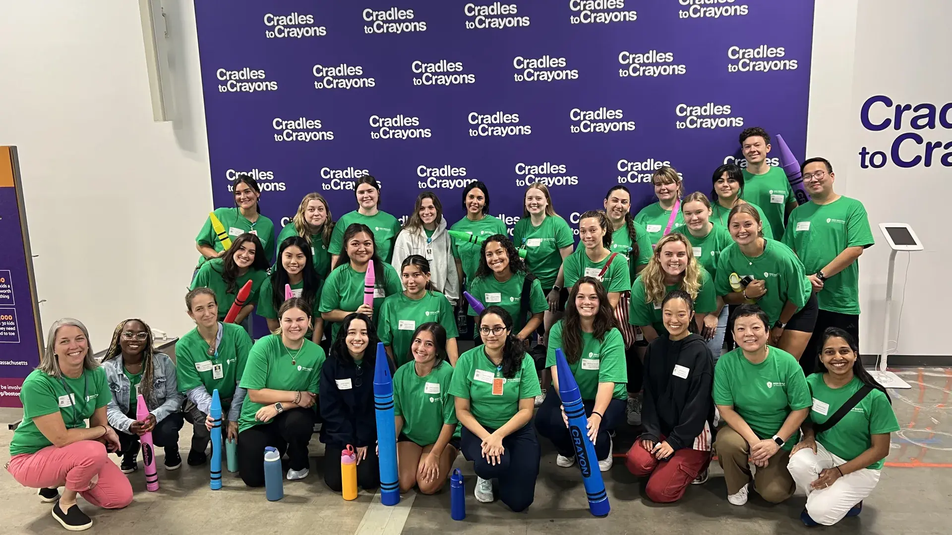 A group of people pose in front of a purple backdrop with the name Cradles to Crayons on it