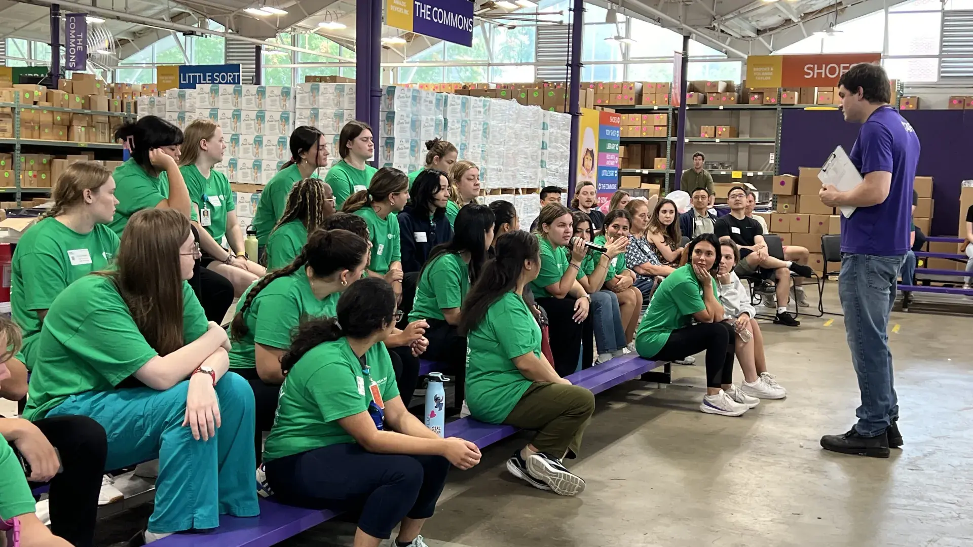 A group of people sit on benches while a person with a clip board tallks to them