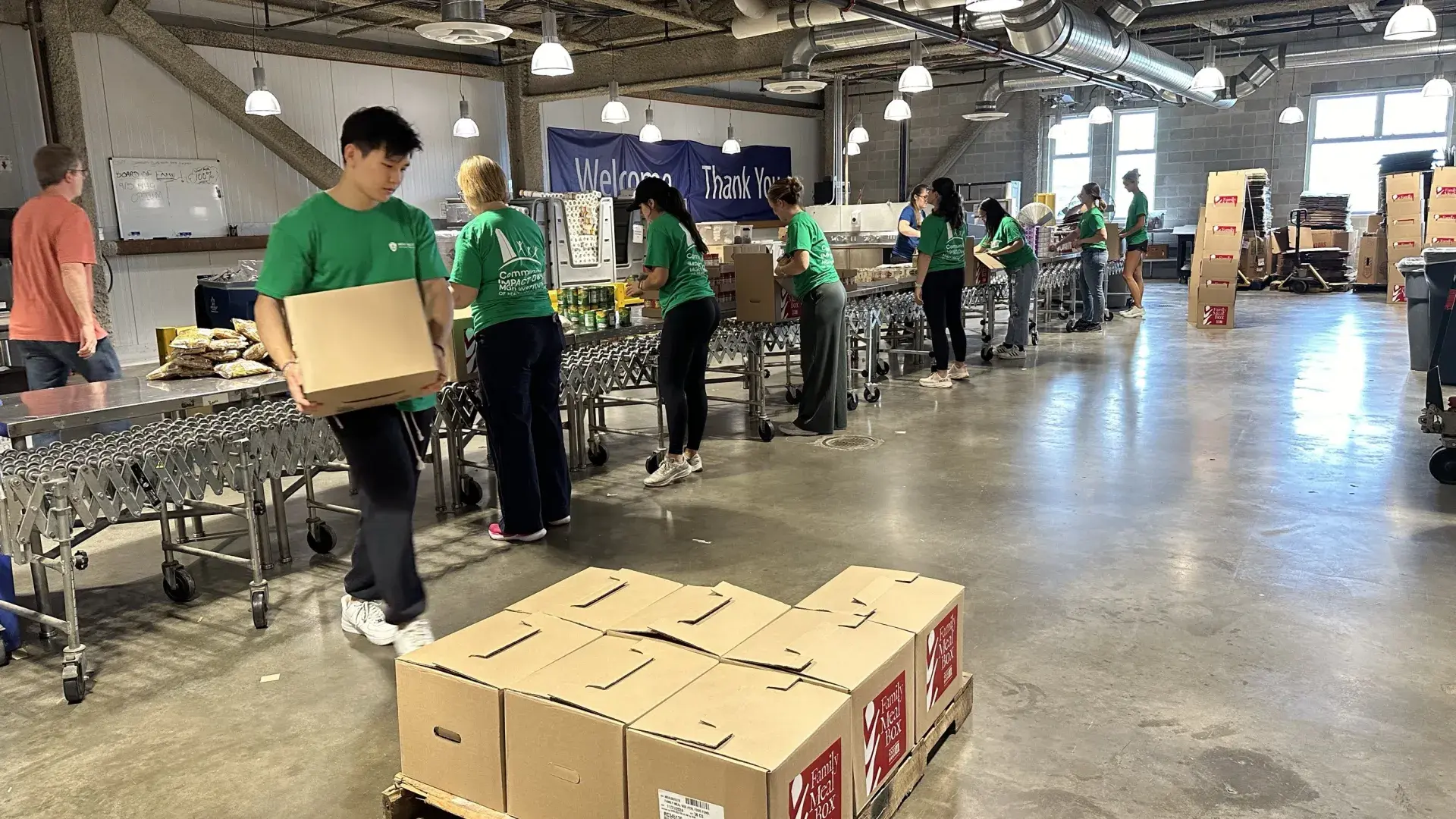 People work at an assembly line filling boxes while a man carries a finished box to a pallet with other boxes