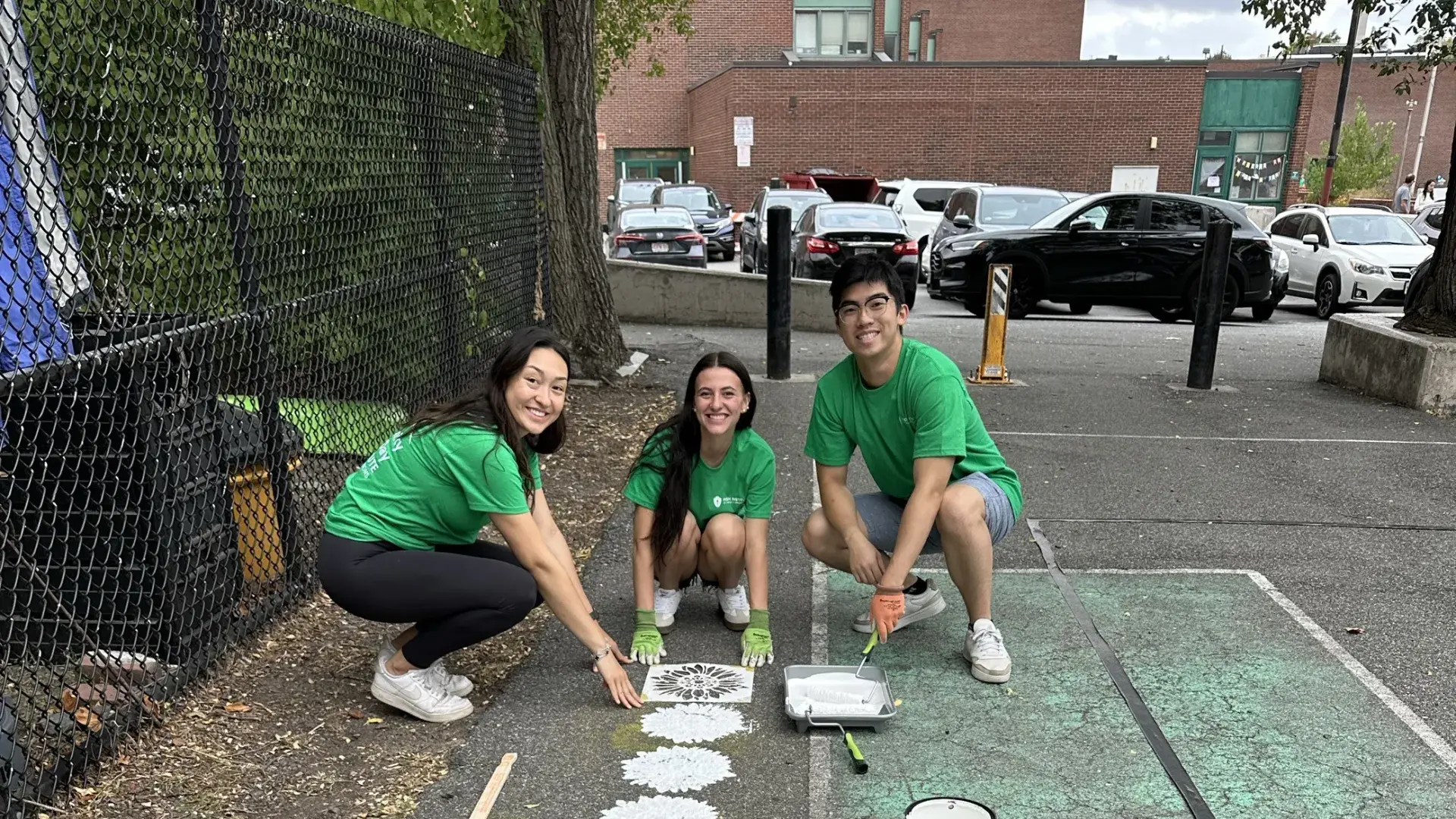 Three people look at the camera while painting flower stencils on the ground of a playground