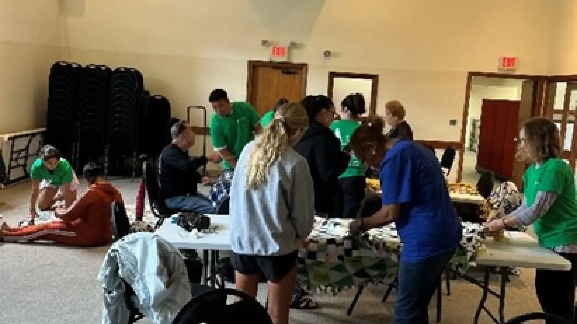 There are rolled up blankets on a table in the foreground while people work with blanks at tables behind it