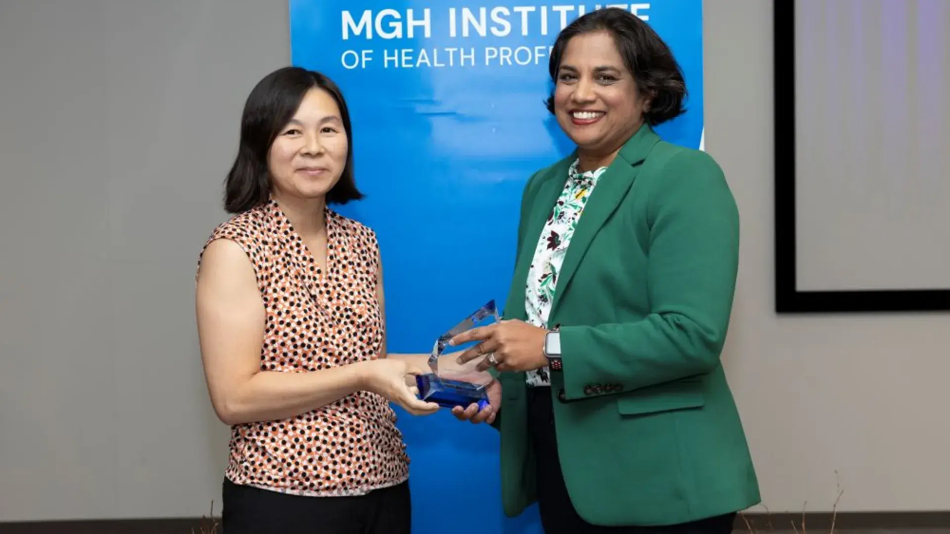 two women stand in front of back drop that says MGH institute holding an award