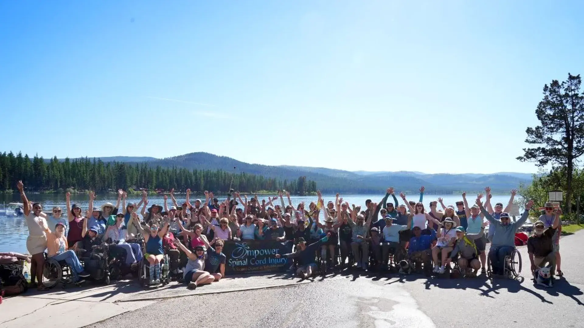 A large group of people stand together for a photograph in front of a lake with trees and mountains in the background