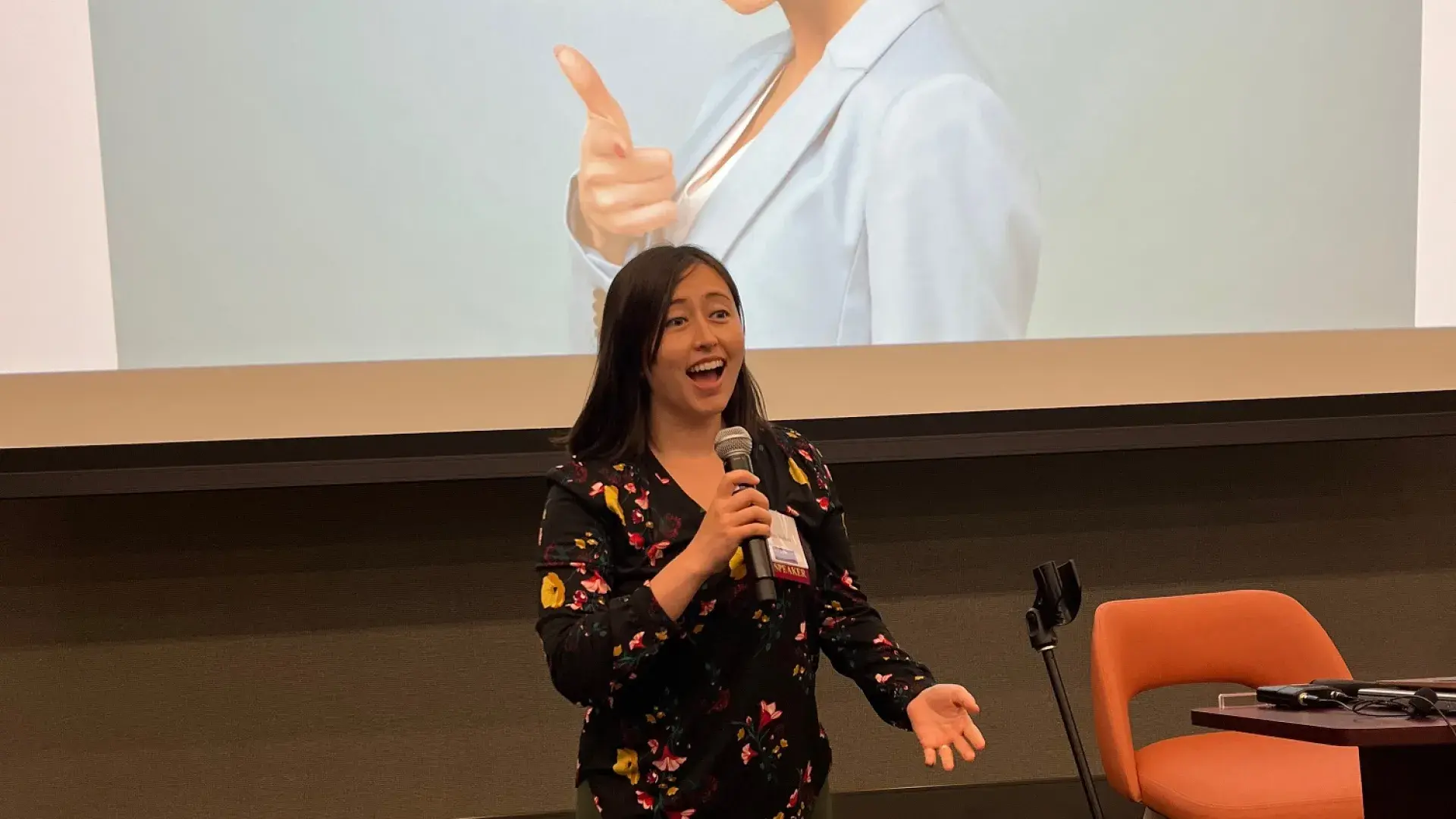 A woman stands in front of a screen holding a microphone