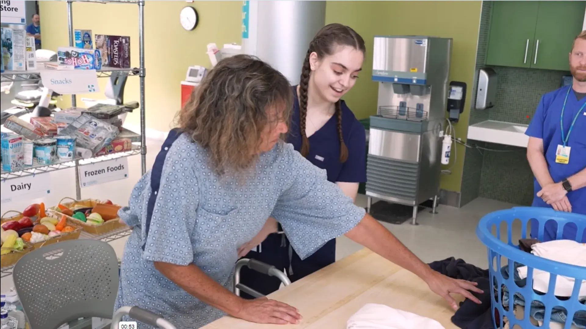 A student wearing hospital scrubs helps a woman wearing a hospital gown practice folding the laundry while a man with an id bade watches