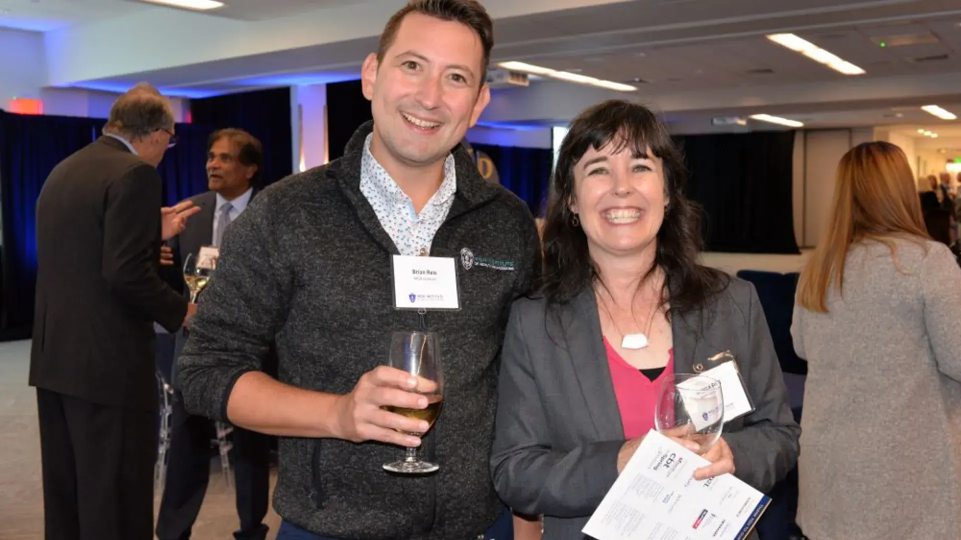Two smiling attendees holding drinks and event materials at the MGH Institute of Health Professions healthcare networking event.