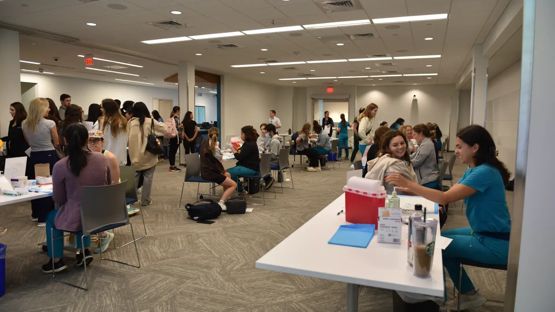 People sit at multiple tables with their arms ready to get a flu shot while nursing students administer them and a crowd waits on the side of the room 