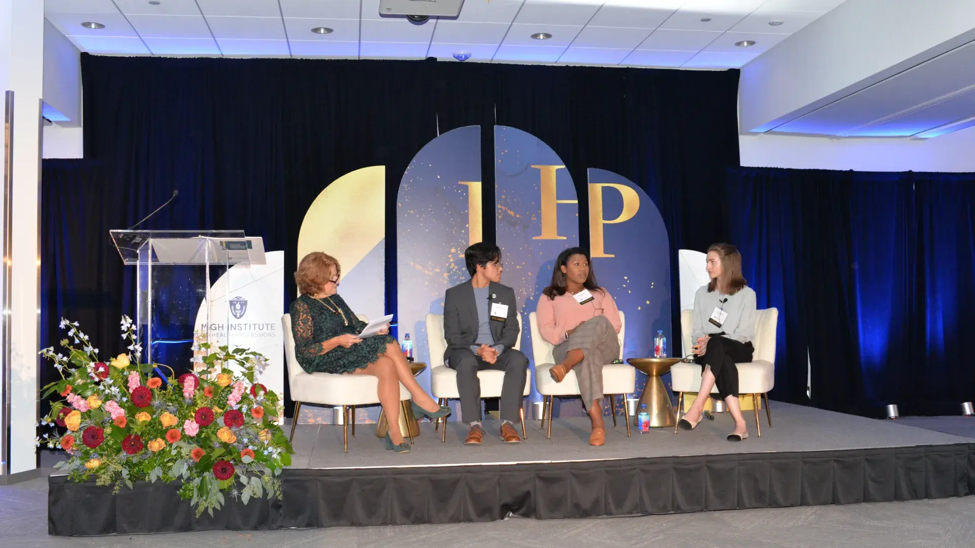 Three women sit in chairs on a stage with a backdrop displaying IHP