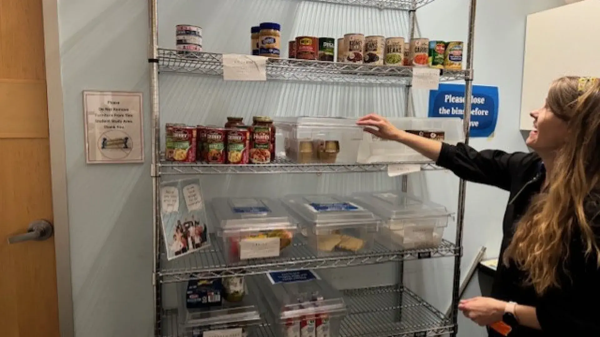 A woman stands in front of metal shelves with cans and boxes of food