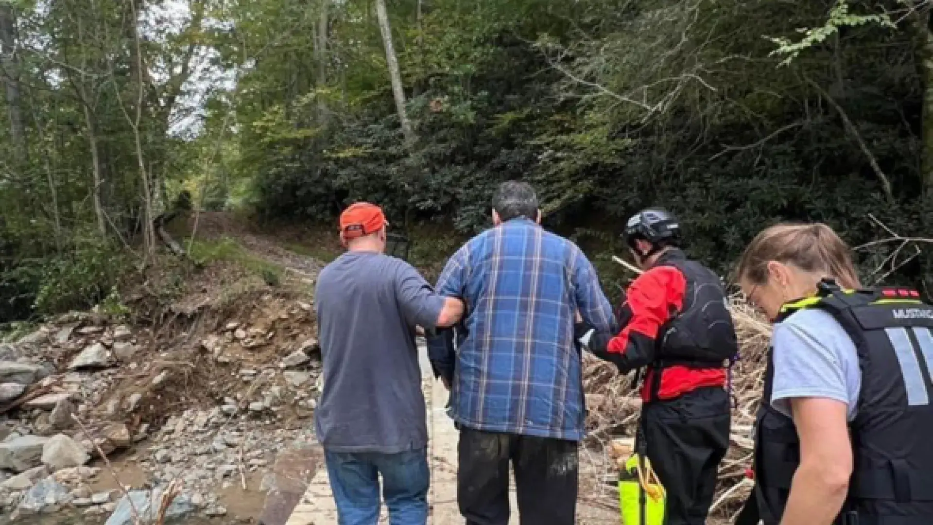 Two people help a third over a path over water while a fourth person trails behinds