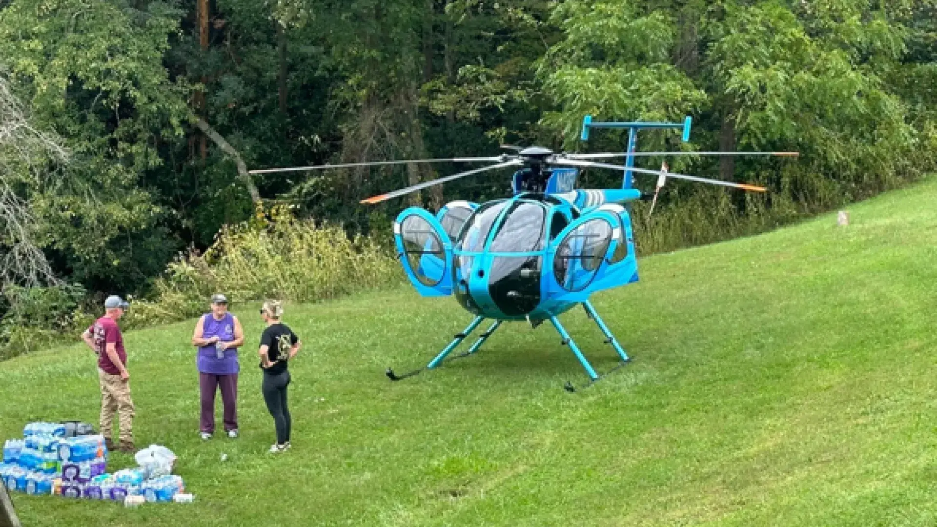 A helicopter sits on a grassy spot while three people stand near supplies