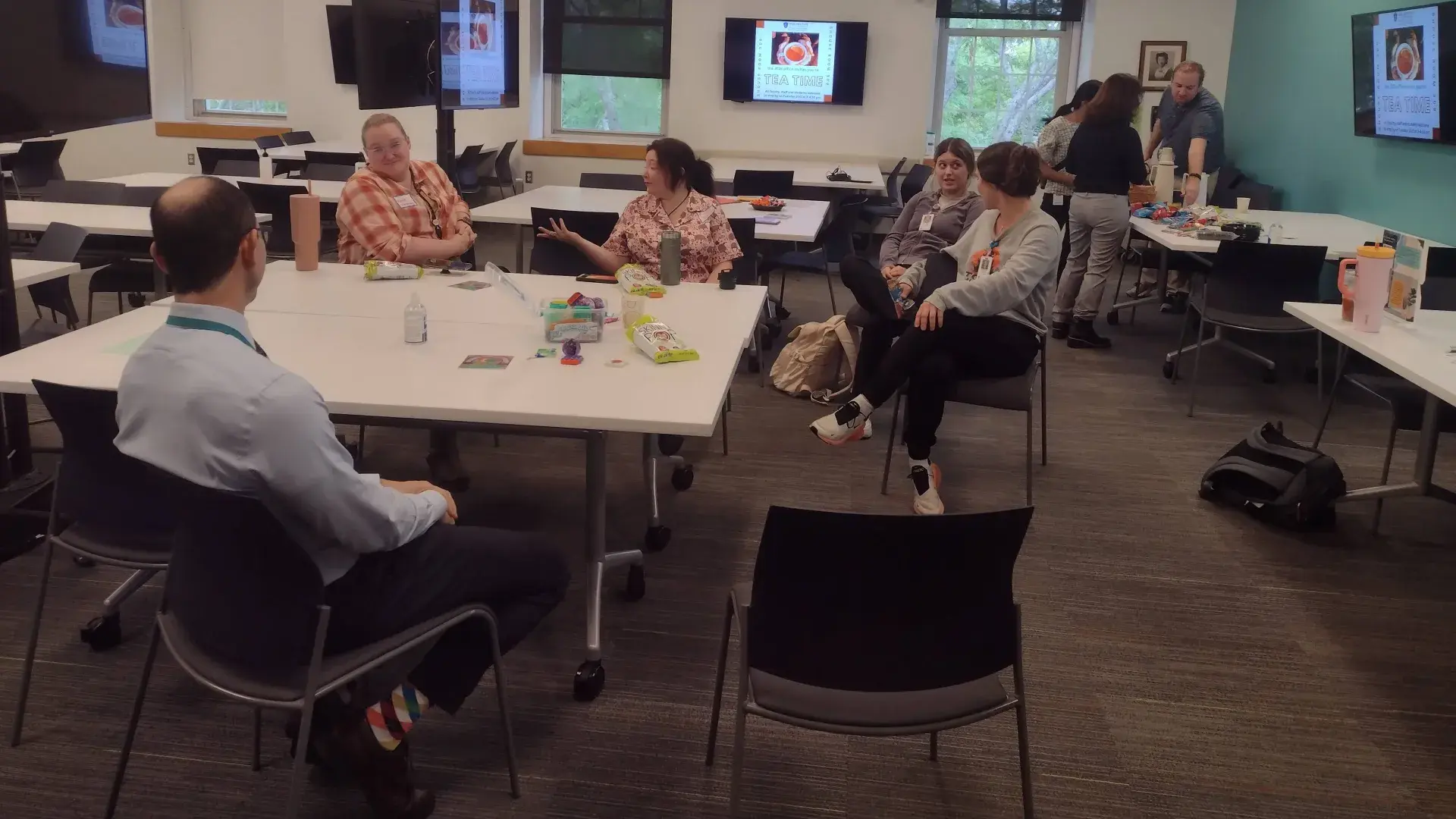 Five people sit around a table while several people in the background stand in front of a table being served food and drinks