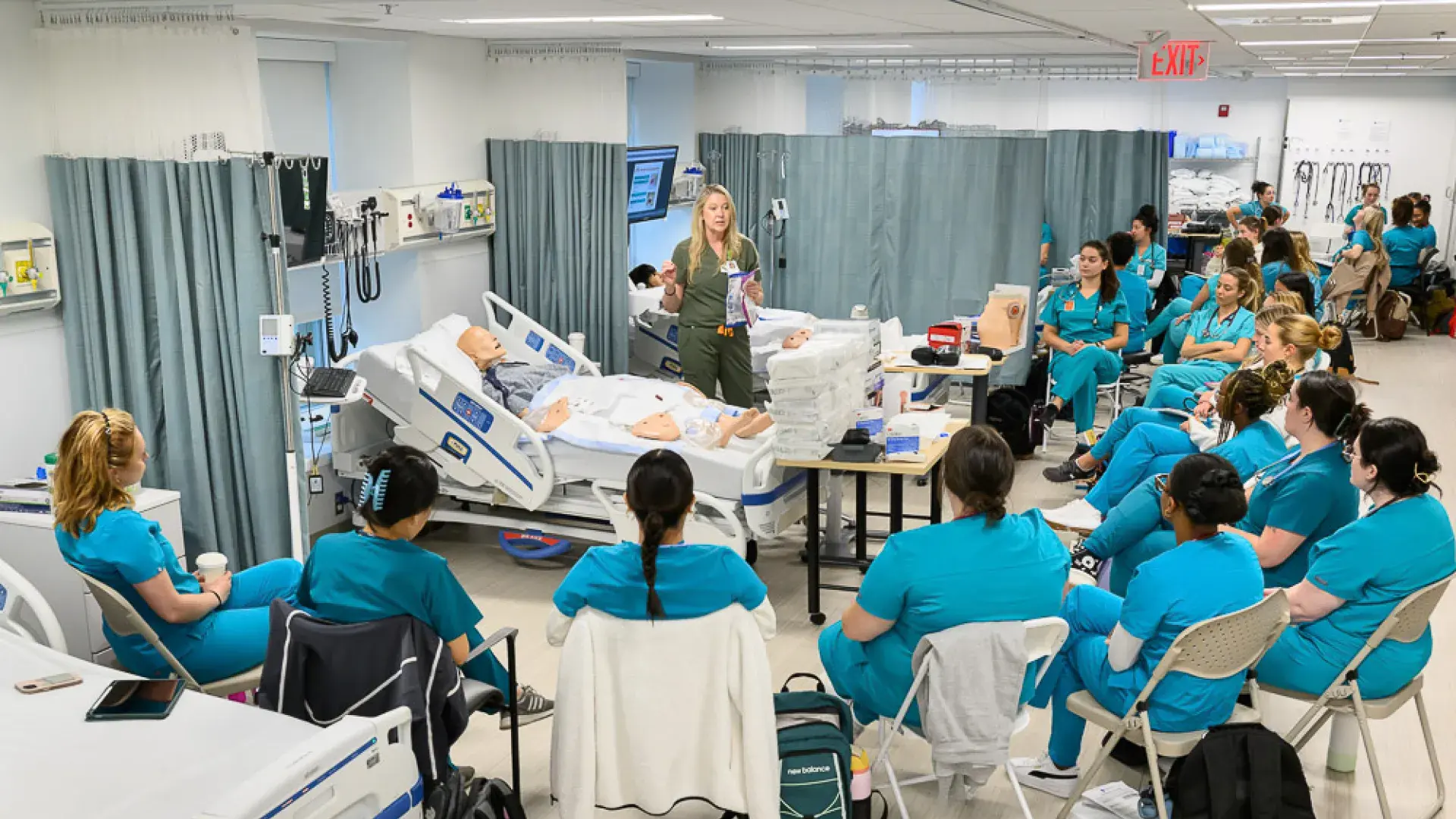a woman stands in front of a manikin on a hospital bed while people in scrubs sit in a semi circle around her