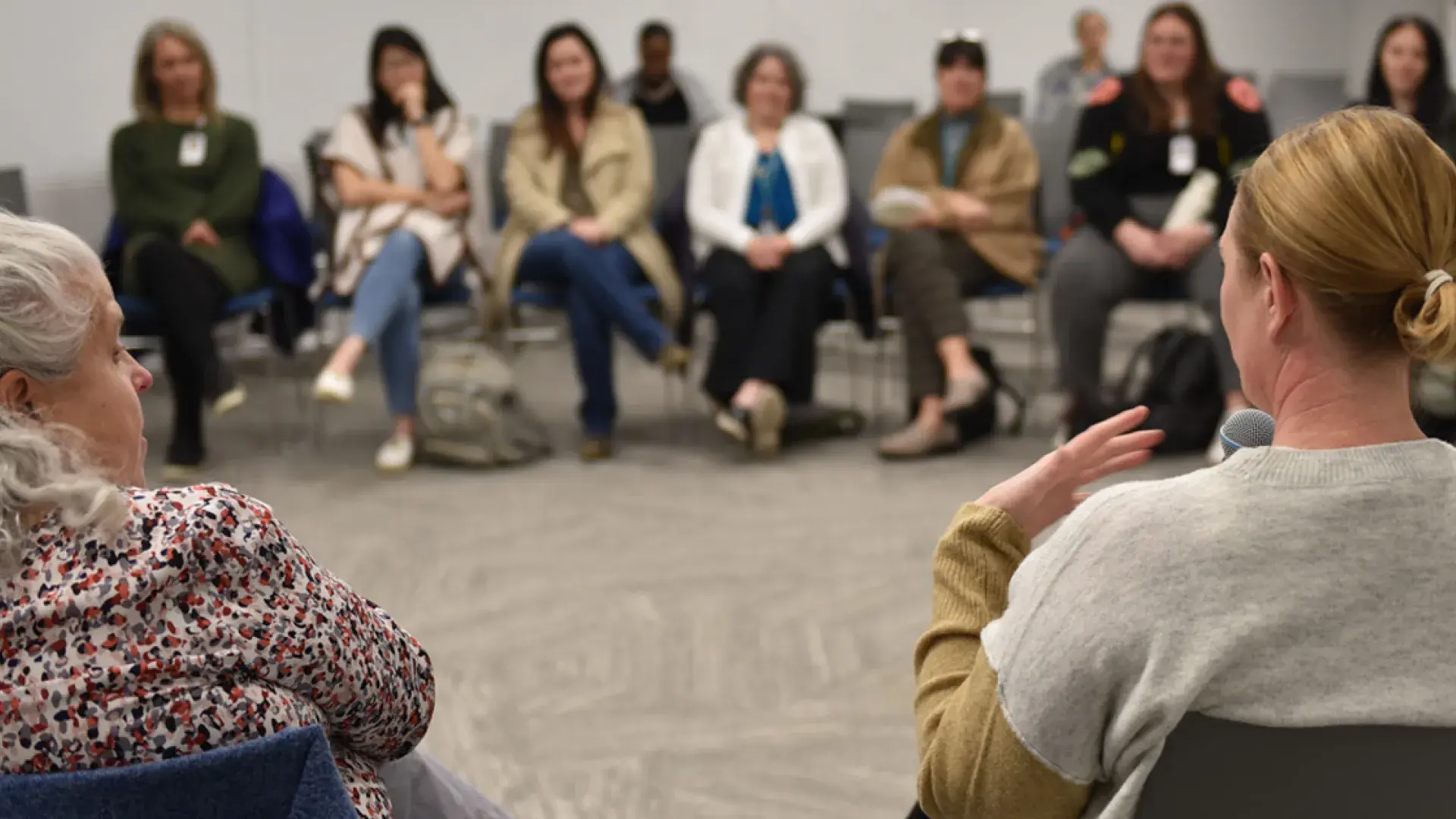 A woman holding a microphone is shown from behind while a group of people sit across from her listening