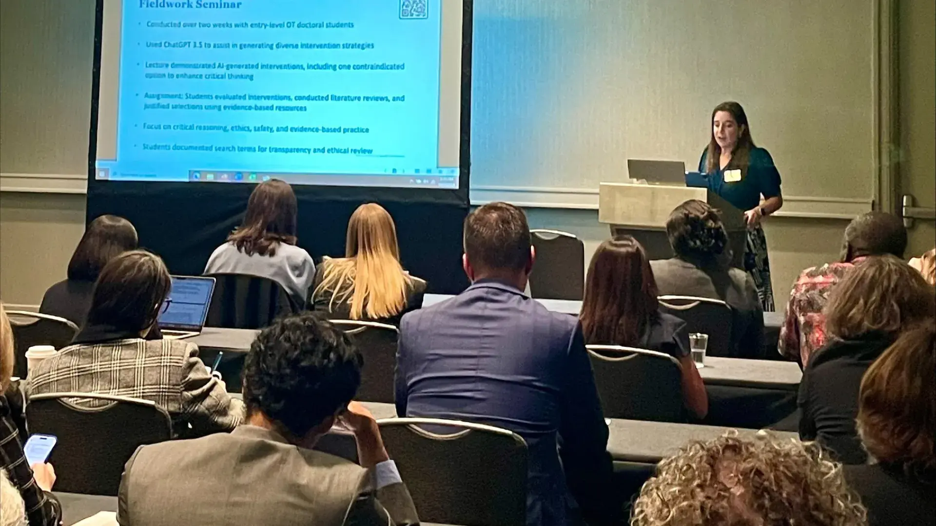A woman stands at a podium with a screen to her right and an audience sitting in front of her