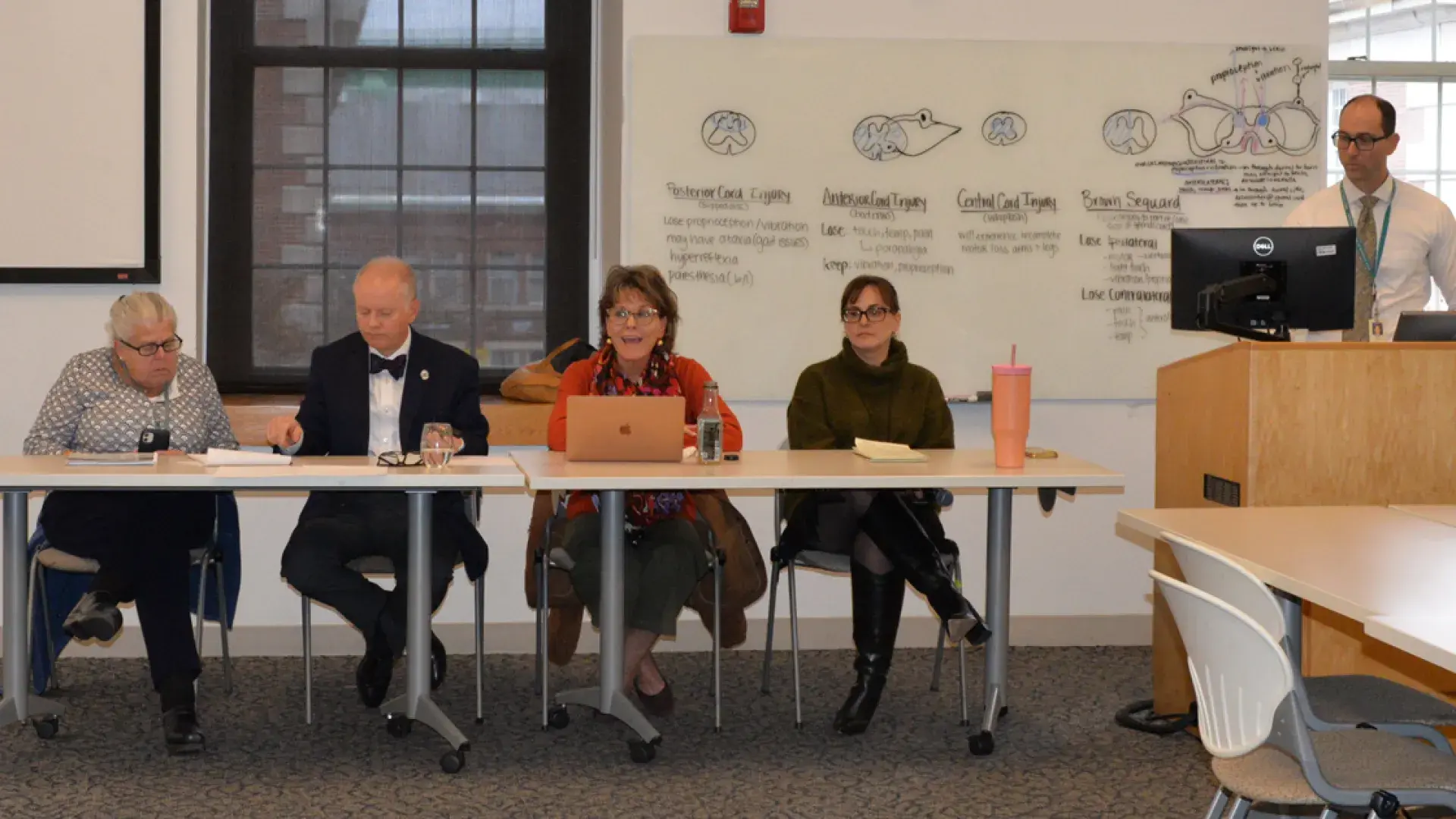 four people sit behind a table while a fifth person is at a podium with a computer