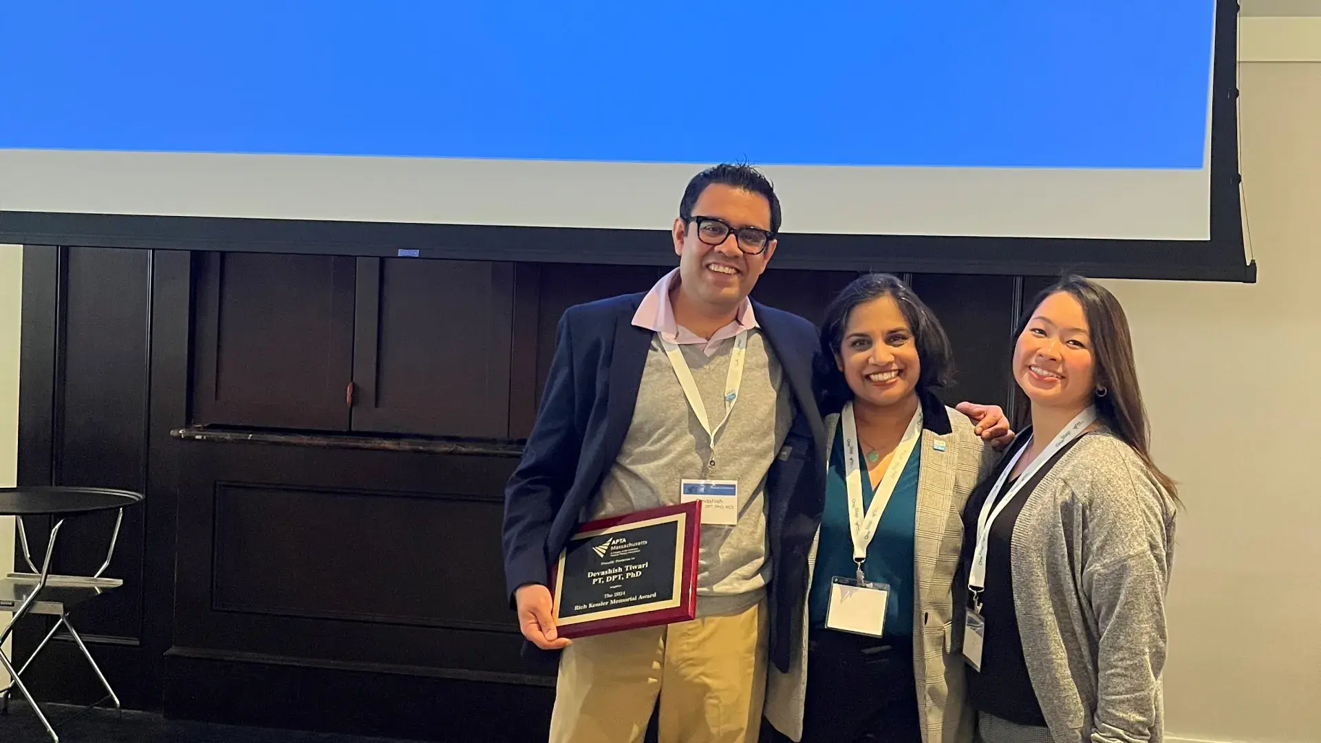 Three people stand in front of a screen with the words APTA Massachusetts and one is holding a plaque