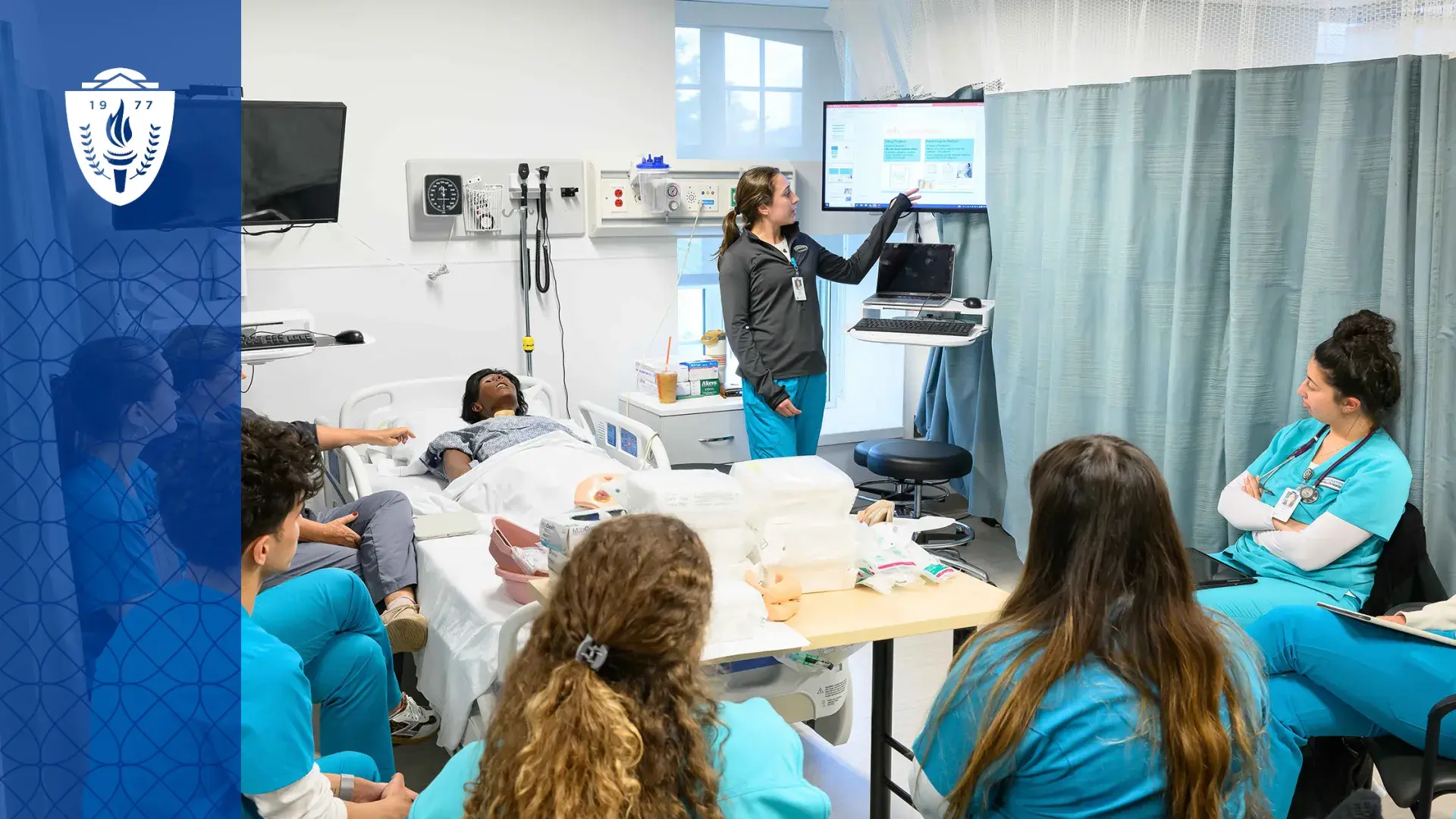 students in scrubs sit around a hospital bed containing a manikin while a woman points at a monitor above the bed