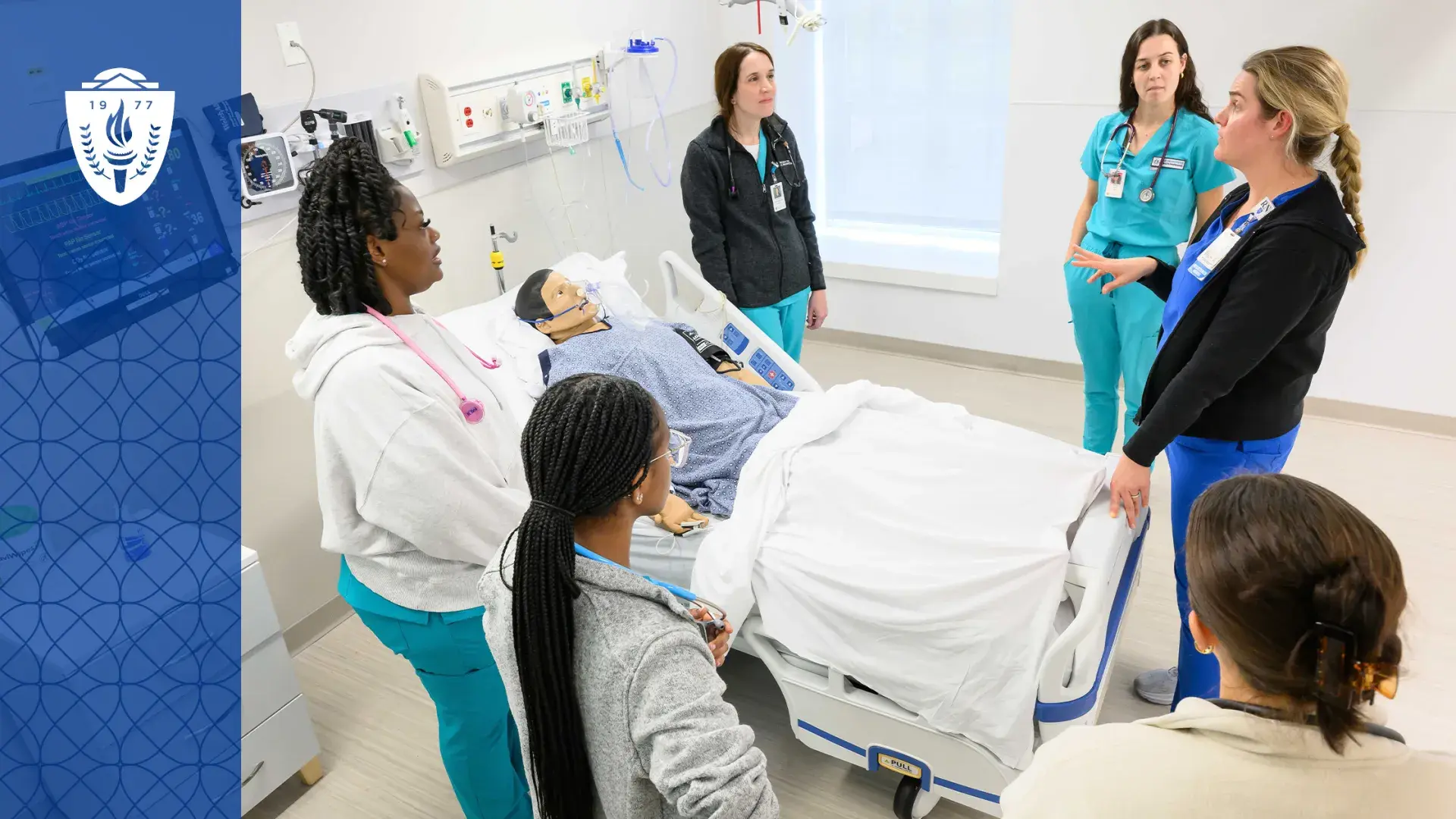 Education in the Health Professions is changing, here we see a faculty member in scrubs surrounded by five students in scrubs around a hospital bed with a manikin