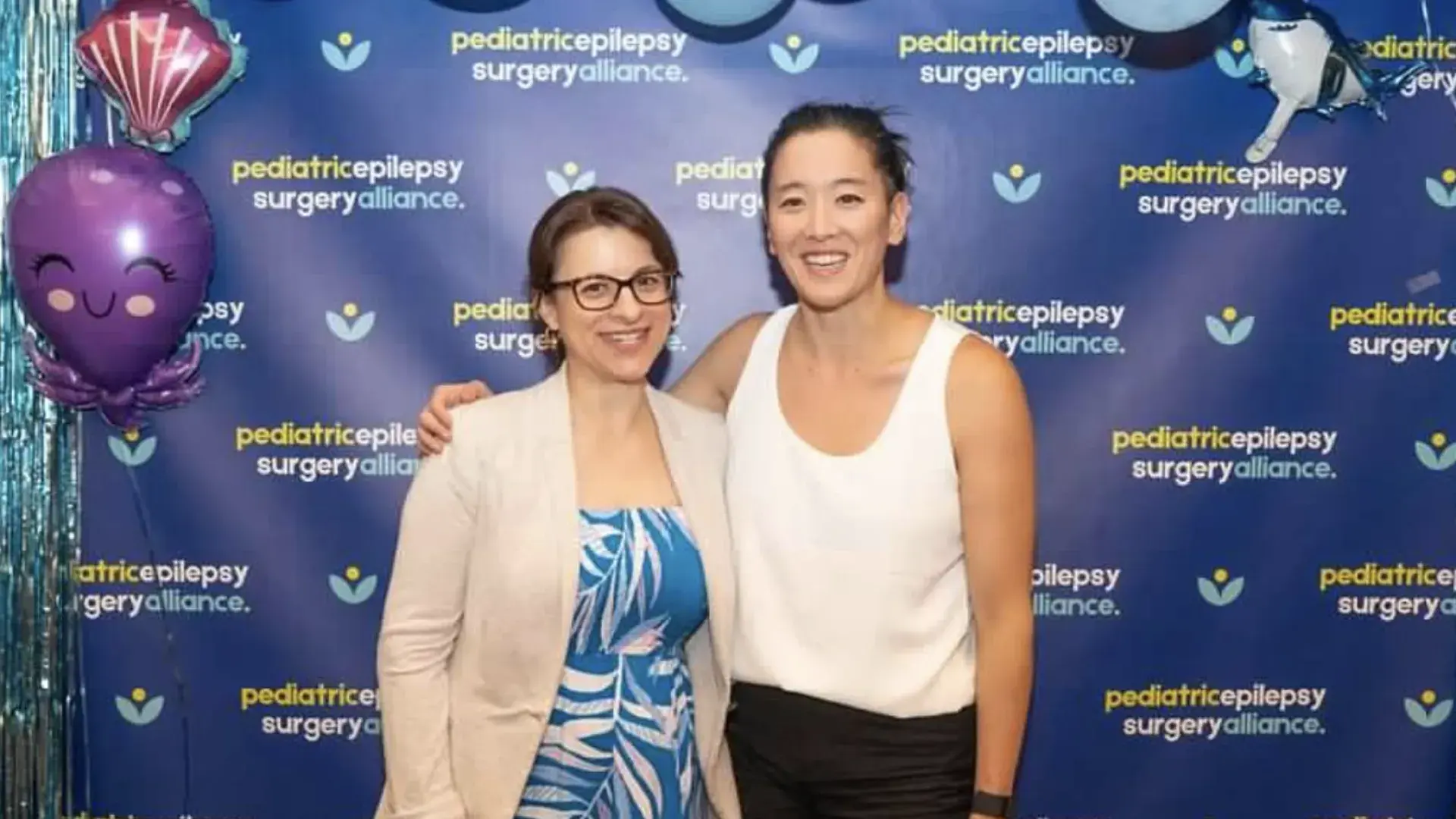 Two women stand in front of a back drop