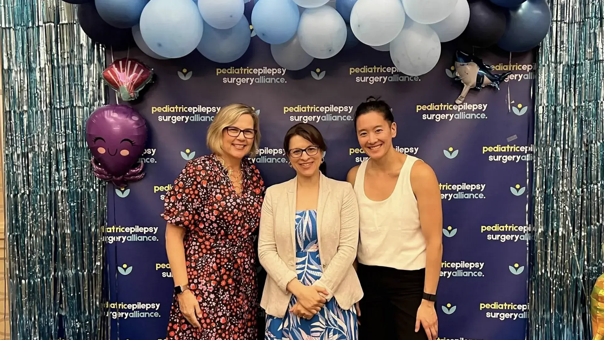 Three women stand in front of a back drop with balloons overhead