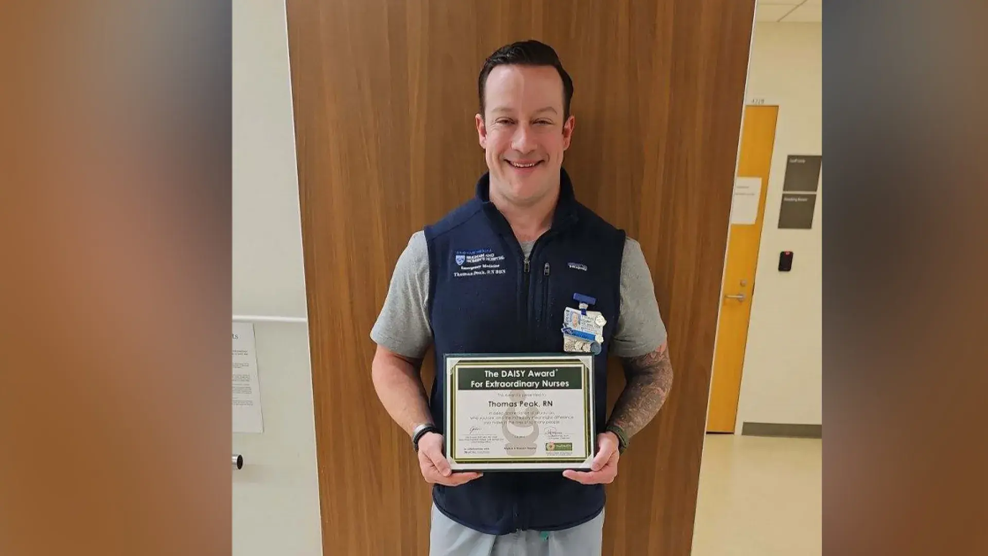A man stands with his back to a wall holding a certificate for the Daisy Award for Extraordinary Nurses