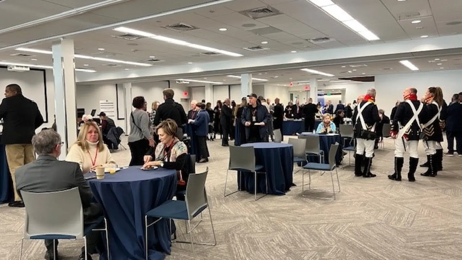 People at an event sit in chairs around tables while others stand in groups talking