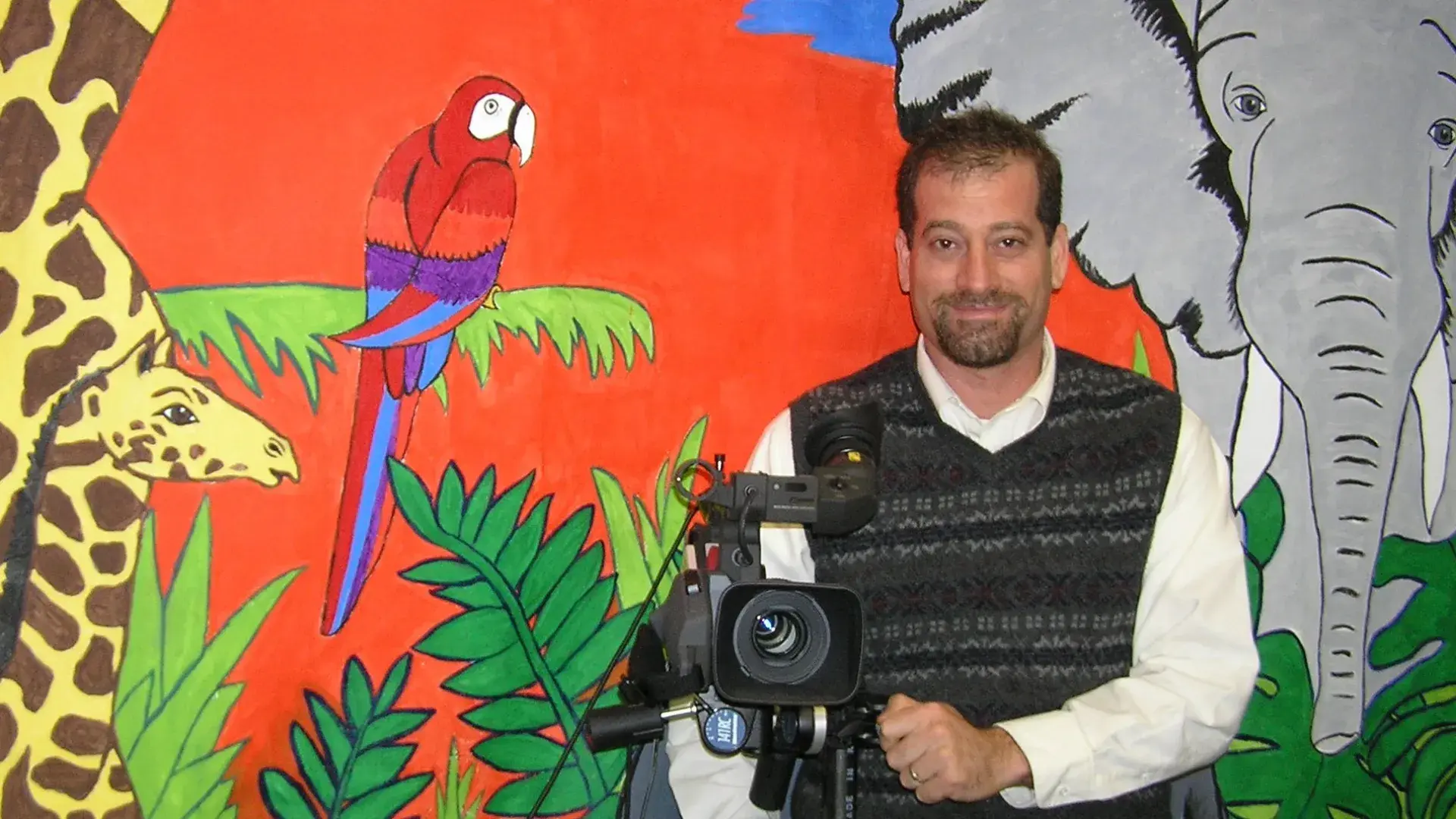 A man stands in front of a colorful wall with giraffes, a parrot and elephant painted on it. 