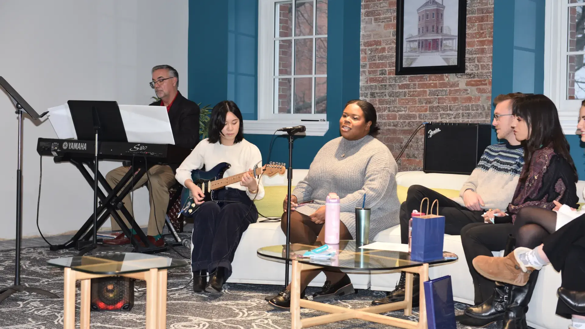 Four people sit on a couch, one has a guitar and a man sits behind a keyboard to their right