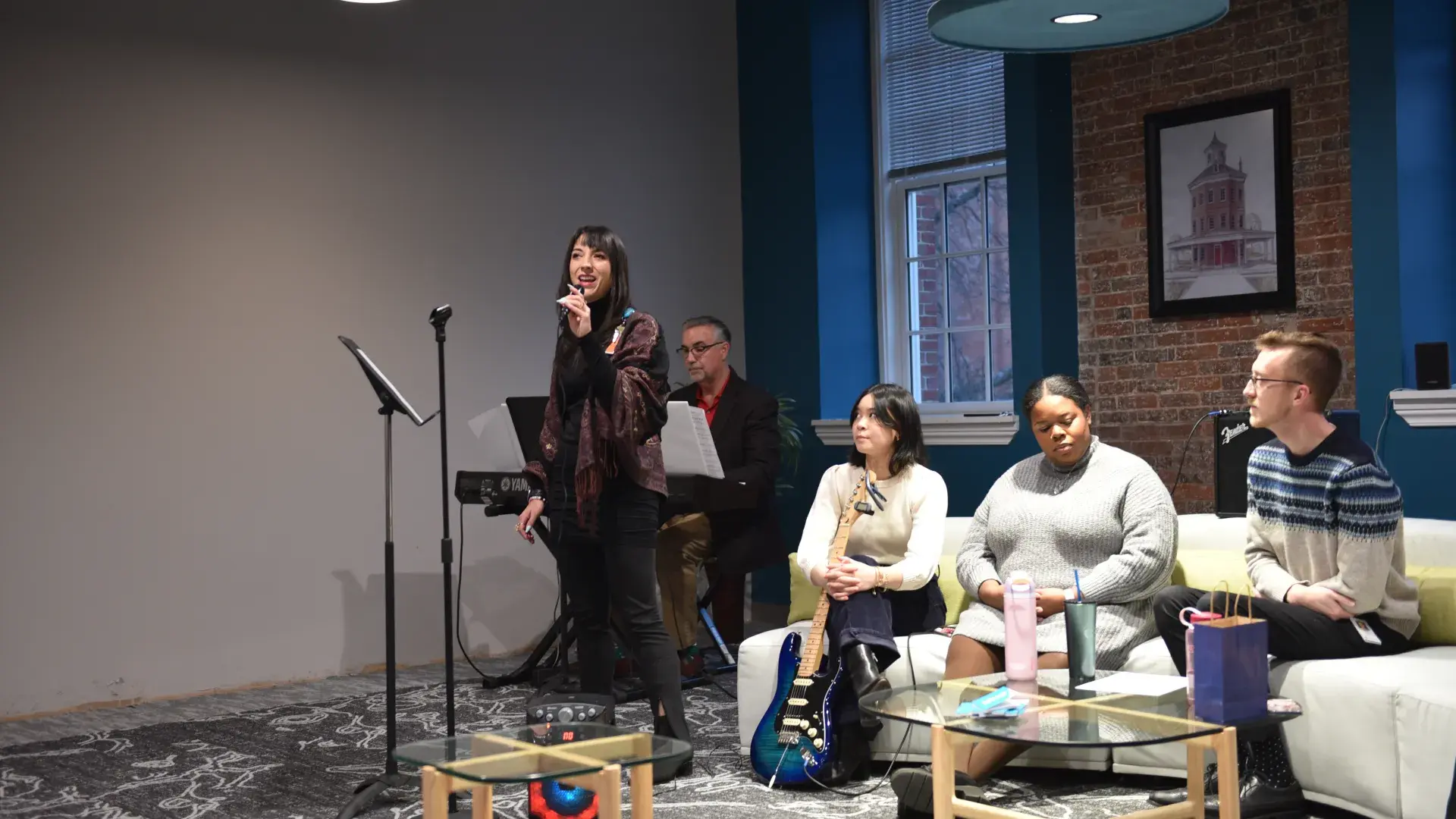 A woman sings into a microphone while a man plays the keyboard behind her and a woman holding a guitar sits on a couch. 