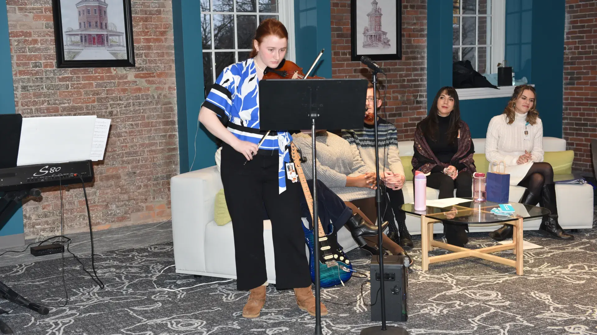 A woman plays the violine while people look on from a couch behind her