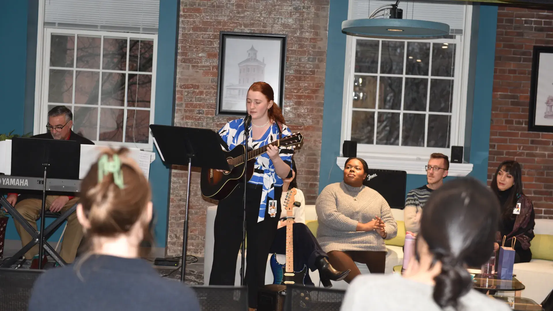 A woman plays the guitar while a man plays the keyboard behind her and four people sit on coach watching from behind her