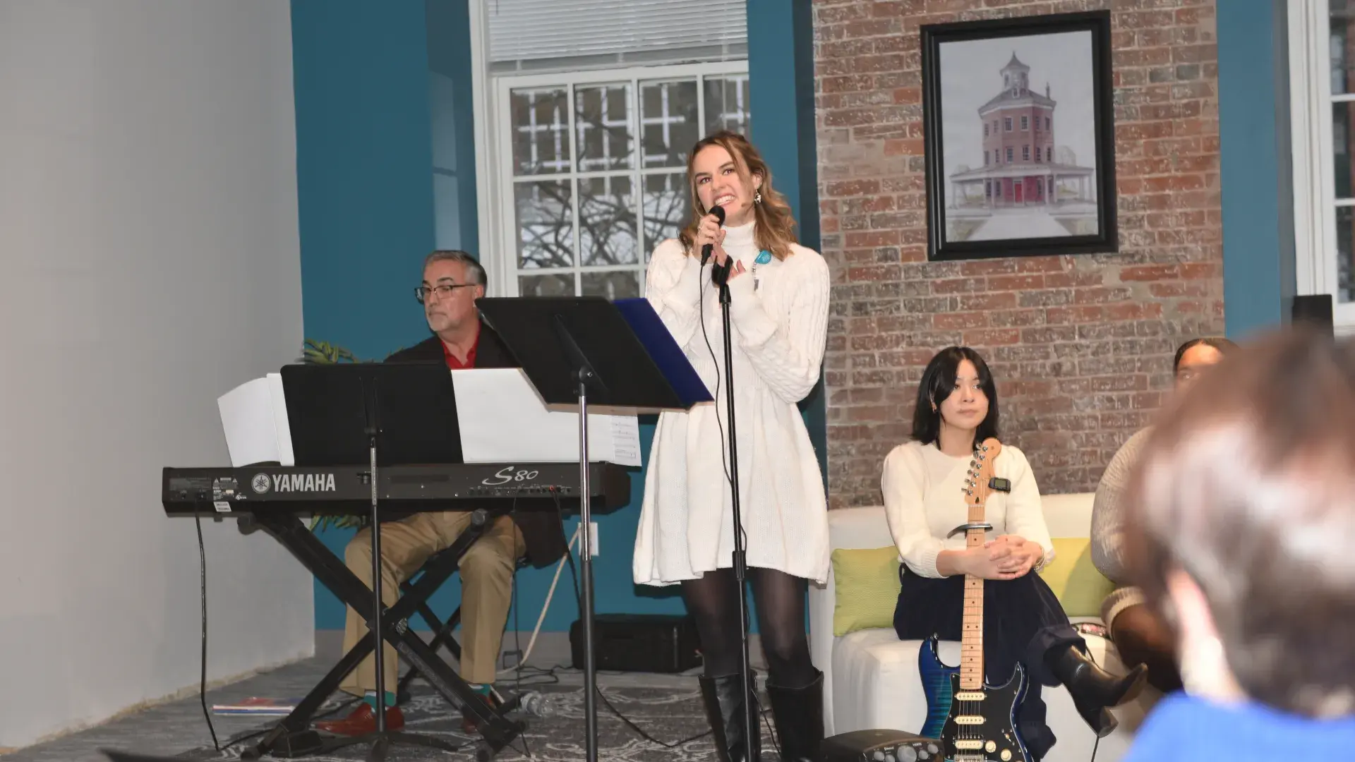A woman stands behind a microphone while a man sits behind the keyboard to her right and a woman holds a guitar while sitting on a couch to her left