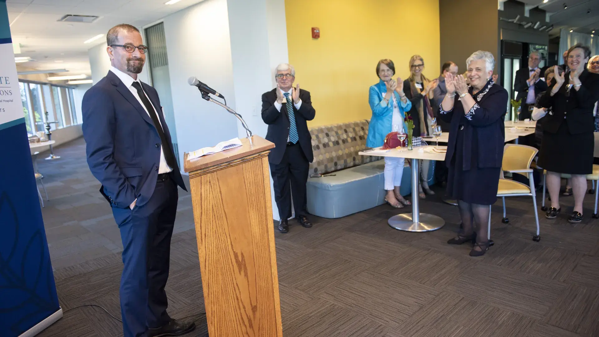 A man stands at a podium on the left while people stand and clap