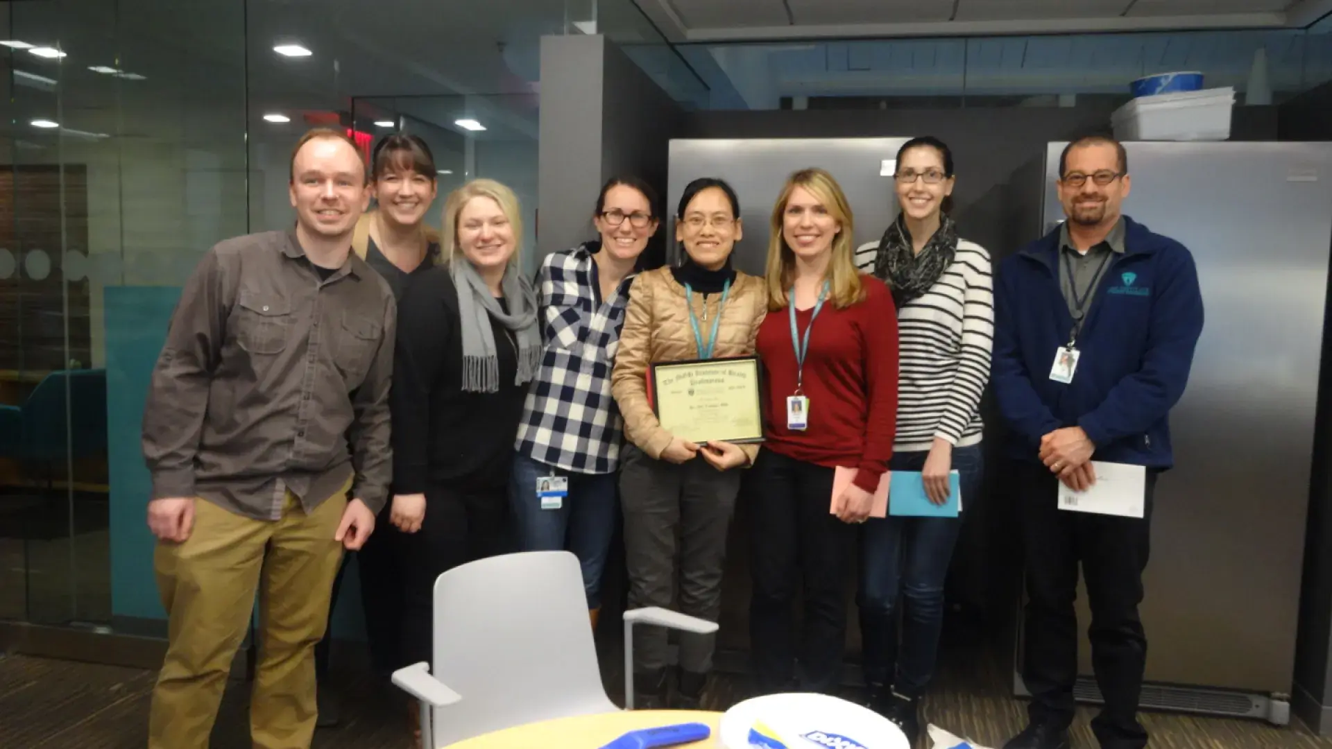 Nine people pose in an office with the person in the middle holding a certificate