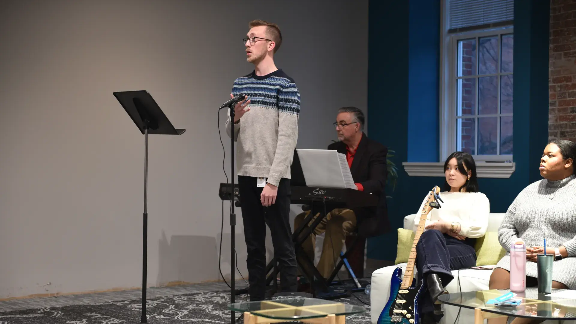 A man stands behind the microphone while another man sits behind keyboards and two women sit on a couch behind him 