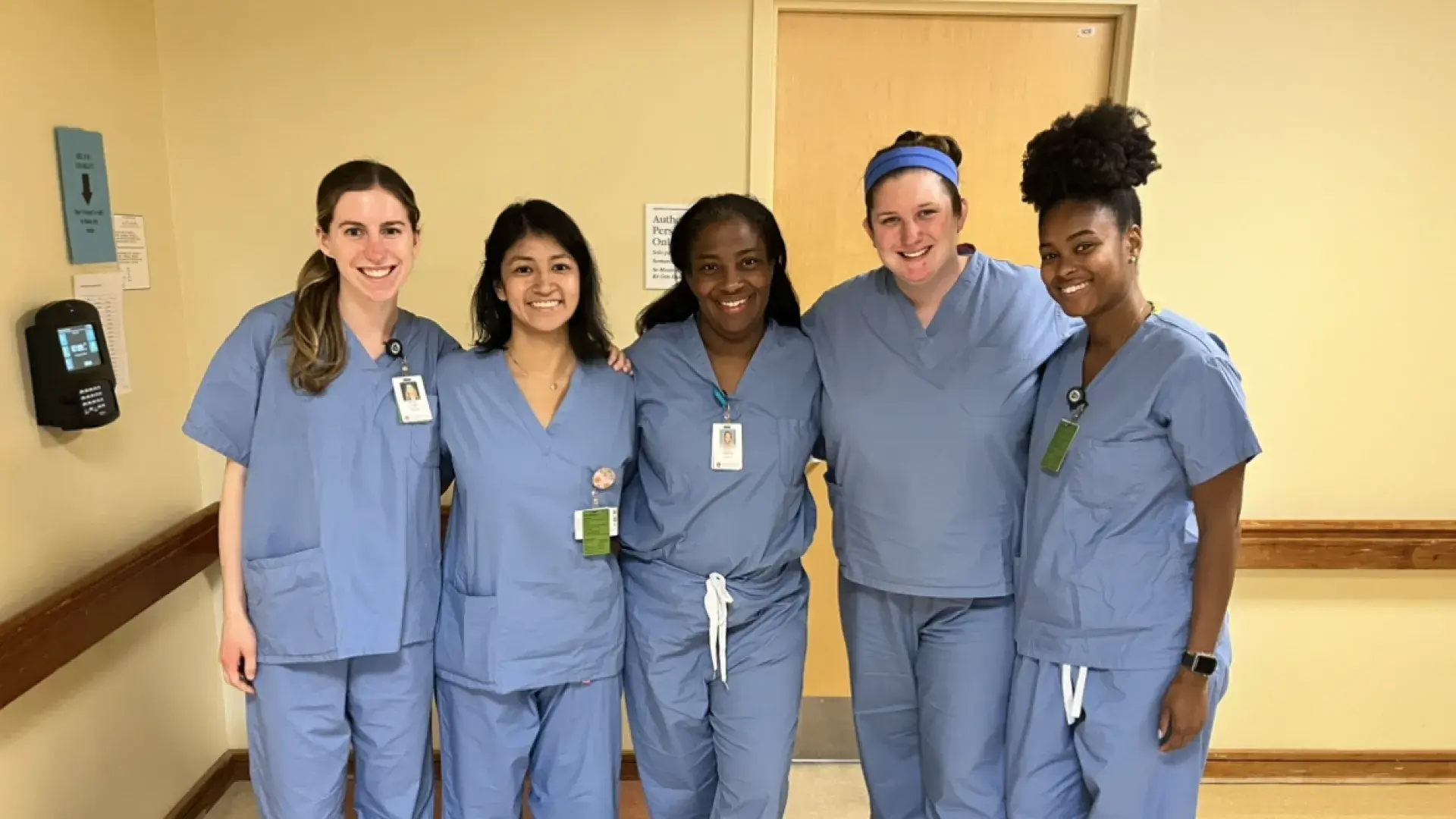 Five women wearing medical scrubs stand in a hallway posing for the camera