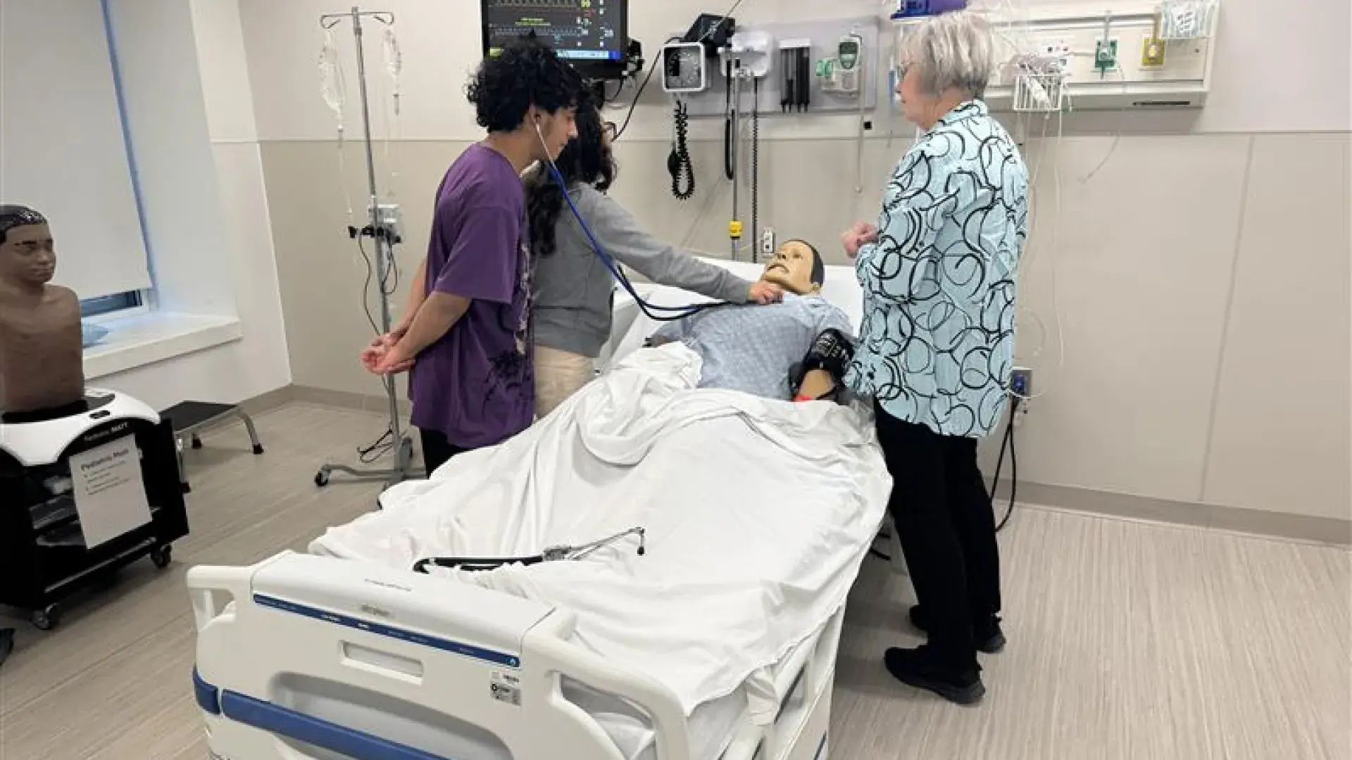 A person holds a stethoscope to a mannikin while another listens to the other end of the stethoscope while a third person looks on. 