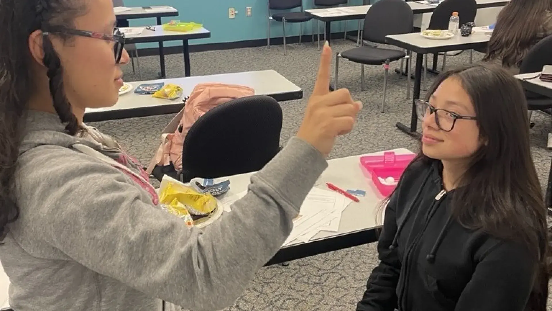 A woman with the finger of her right hand held up stands in front of a woman who is sitting and watching the finger